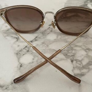 May include: Cat-eye sunglasses with a brown gradient lens and a gold-tone metal frame. The temples are thin and gold, with brown tips. The sunglasses are set against a marble background.