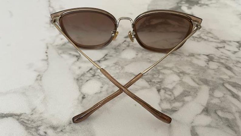 May include: Cat-eye sunglasses with a brown gradient lens and a gold-tone metal frame. The temples are thin and gold, with brown tips. The sunglasses are set against a marble background.