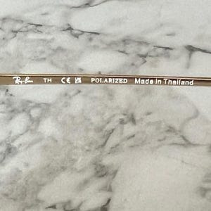 May include: Close-up of the temple of a pair of gold-toned sunglasses. The text "Ray-Ban", "POLARIZED", and "Made in Thailand" are visible. The glasses have a sleek, modern design.