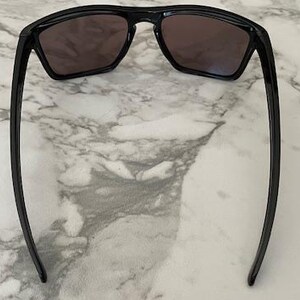 May include: Black sunglasses with dark lenses are displayed against a marble background. The sunglasses have a rectangular frame and long, slender arms. The lenses appear to be tinted, offering protection from the sun.