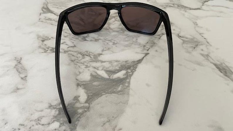 May include: Black sunglasses with dark lenses are displayed against a marble background. The sunglasses have a rectangular frame and long, slender arms. The lenses appear to be tinted, offering protection from the sun.