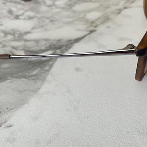 May include: A pair of sunglasses with brown frames and temples. The temples are a silver color and have brown tips. The sunglasses are resting on a white and gray surface.
