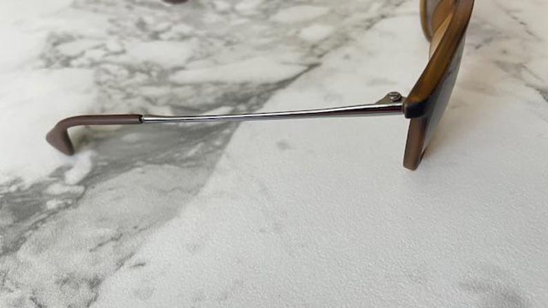 May include: A pair of sunglasses with brown frames and temples. The temples are a silver color and have brown tips. The sunglasses are resting on a white and gray surface.
