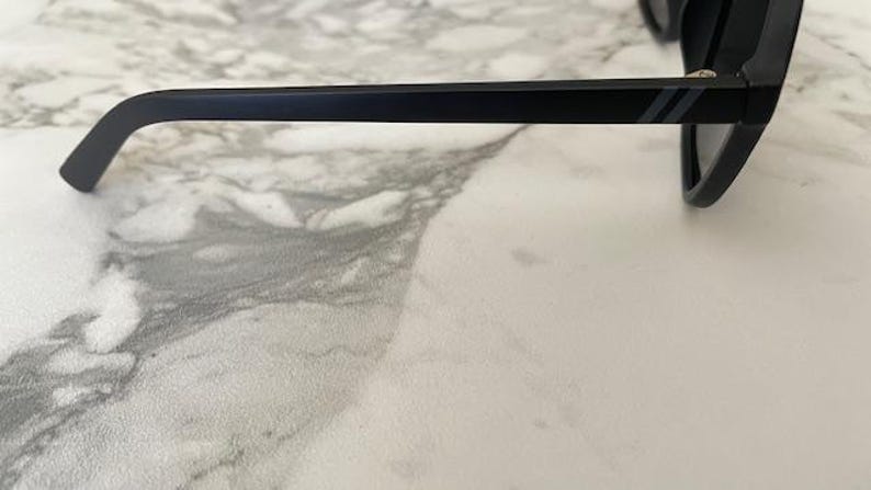 Puede incluir: Un par de gafas de sol negro mate con una patilla recta que presenta dos finas rayas diagonales grises paralelas cerca de la bisagra. La montura est&aacute; fabricada con un material negro s&oacute;lido con lentes oscuras visibles en el lado derecho.