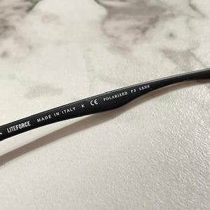 May include: Close-up of a black Ray-Ban Liteforce eyeglass arm. The arm has the Ray-Ban logo, the text "LITEFORCE", "MADE IN ITALY", and "POLARIZED P3 LENS" printed on it. The arm is resting on a light-colored surface.