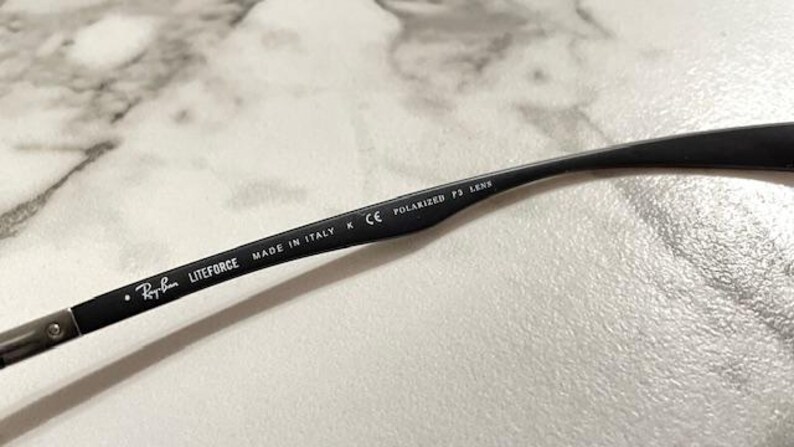 May include: Close-up of a black Ray-Ban Liteforce eyeglass arm. The arm has the Ray-Ban logo, the text "LITEFORCE", "MADE IN ITALY", and "POLARIZED P3 LENS" printed on it. The arm is resting on a light-colored surface.