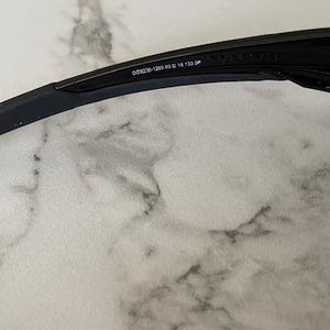 May include: Black sunglasses with a sleek, modern design. The frame is black with a curved arm and a rectangular lens. The text "OO9326-1260 53 16 133 JP" is printed on the arm.
