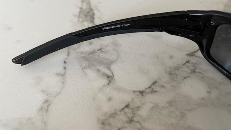 May include: Black sunglasses with a sleek, modern design. The frame is black with a curved arm and a rectangular lens. The text "OO9326-1260 53 16 133 JP" is printed on the arm.