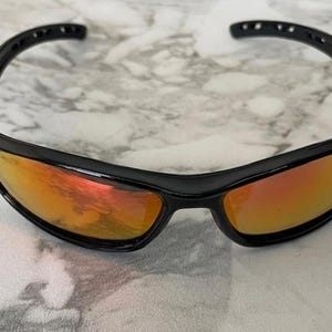 May include: Black sport sunglasses with orange mirrored lenses. The sunglasses have a sleek, curved design with a black frame. The lenses reflect a warm, orange hue, offering a stylish and functional accessory for outdoor activities.