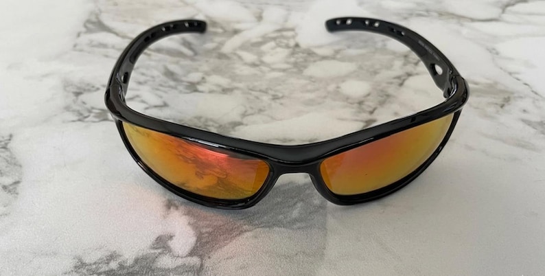 May include: Black sport sunglasses with orange mirrored lenses. The sunglasses have a sleek, curved design with a black frame. The lenses reflect a warm, orange hue, offering a stylish and functional accessory for outdoor activities.