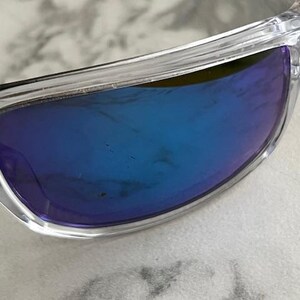 May include: Clear-framed sunglasses with blue-tinted lenses. The lenses have a gradient effect, transitioning from a darker blue at the top to a lighter shade towards the bottom. The frame is transparent, allowing a view of the underlying surface.