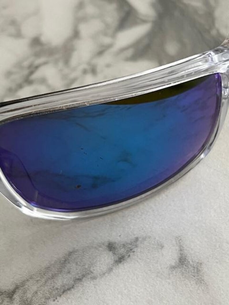 May include: Clear-framed sunglasses with blue-tinted lenses. The lenses have a gradient effect, transitioning from a darker blue at the top to a lighter shade towards the bottom. The frame is transparent, allowing a view of the underlying surface.