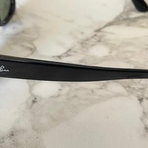 Puede incluir: Gafas de sol negras con lentes verdes. El logotipo de Ray-Ban es visible en la patilla. Las gafas de sol est&aacute;n sobre una superficie de m&aacute;rmol blanco y gris.