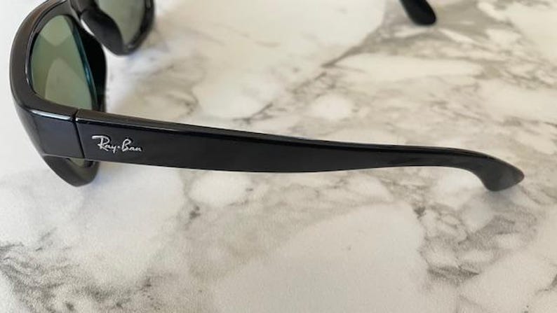 Puede incluir: Gafas de sol negras con lentes verdes. El logotipo de Ray-Ban es visible en la patilla. Las gafas de sol est&aacute;n sobre una superficie de m&aacute;rmol blanco y gris.
