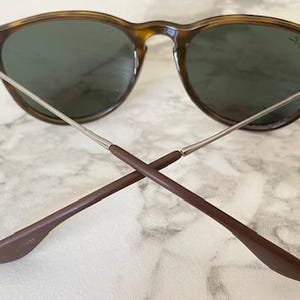 K&ouml;nnte beinhalten: Sonnenbrille mit Schildpatt-Fassung und dunklen Gl&auml;sern. Die B&uuml;gel sind silberfarben mit braunen Spitzen, am unteren Ende gekreuzt. Das Ray-Ban-Logo ist auf dem Glas sichtbar. Auf einem Marmorhintergrund.