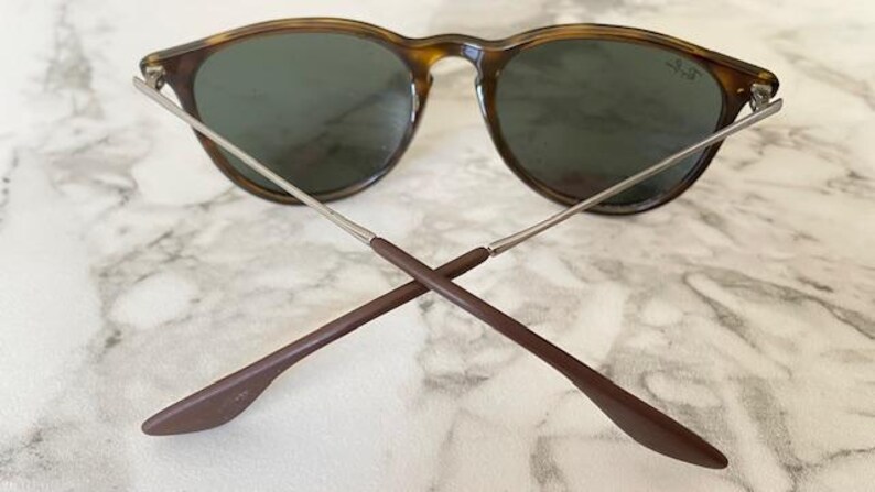 K&ouml;nnte beinhalten: Sonnenbrille mit Schildpatt-Fassung und dunklen Gl&auml;sern. Die B&uuml;gel sind silberfarben mit braunen Spitzen, am unteren Ende gekreuzt. Das Ray-Ban-Logo ist auf dem Glas sichtbar. Auf einem Marmorhintergrund.