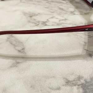 May include: Red sports sunglasses with a sleek, curved design. The sunglasses have a glossy finish and dark lenses. The frame is a deep red color, and the Oakley logo is visible on the temple.
