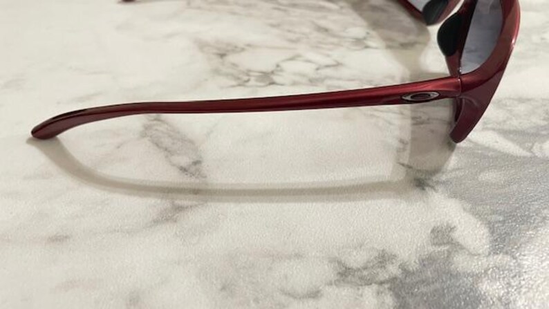 May include: Red sports sunglasses with a sleek, curved design. The sunglasses have a glossy finish and dark lenses. The frame is a deep red color, and the Oakley logo is visible on the temple.