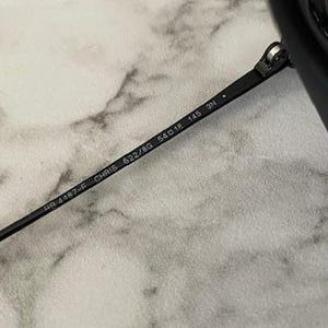 Puede incluir: Primer plano de gafas de sol negras con el texto "RB 4187-F CHRIS 622/8G 54o18 145 3N" impreso en la patilla. Las gafas de sol est&aacute;n sobre un fondo de m&aacute;rmol gris claro y blanco.