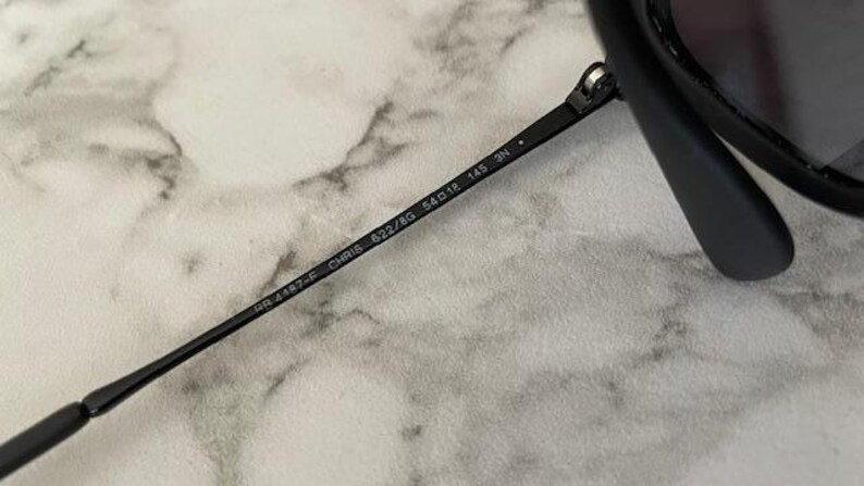 Puede incluir: Primer plano de gafas de sol negras con el texto "RB 4187-F CHRIS 622/8G 54o18 145 3N" impreso en la patilla. Las gafas de sol est&aacute;n sobre un fondo de m&aacute;rmol gris claro y blanco.