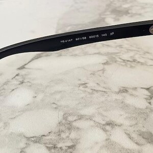 Puede incluir: Gafas de sol negras con lentes oscuras. La patilla es visible, con el texto "RB 4147 601/58 60015 145 3P" impreso. Las gafas est&aacute;n sobre un fondo de m&aacute;rmol.