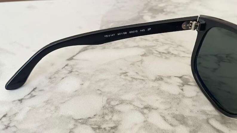 Puede incluir: Gafas de sol negras con lentes oscuras. La patilla es visible, con el texto "RB 4147 601/58 60015 145 3P" impreso. Las gafas est&aacute;n sobre un fondo de m&aacute;rmol.