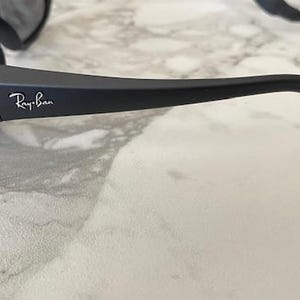 May include: Matte black sunglasses featuring a wrap-around frame design with the white Ray-Ban logo printed on the temple. The sunglasses include dark tinted lenses and a black cord attached to the end of the temple arm for secure placement.