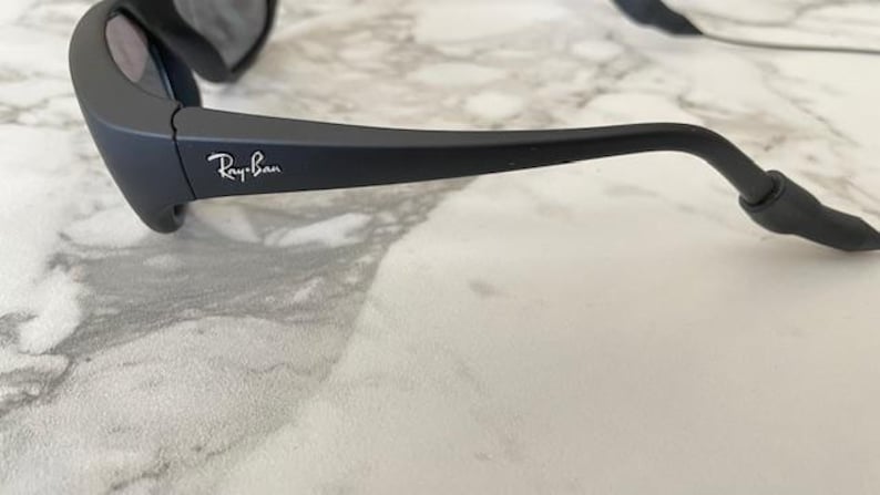 May include: Matte black sunglasses featuring a wrap-around frame design with the white Ray-Ban logo printed on the temple. The sunglasses include dark tinted lenses and a black cord attached to the end of the temple arm for secure placement.