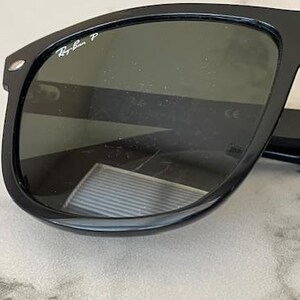 Puede incluir: Gafas de sol negras con lentes oscuras. El logotipo de Ray-Ban es visible en la esquina superior izquierda de la montura. Las gafas de sol tienen una forma rectangular cl&aacute;sica.