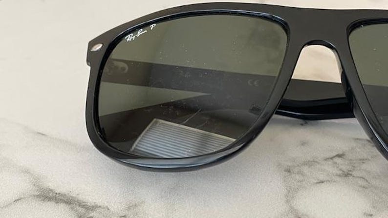 Puede incluir: Gafas de sol negras con lentes oscuras. El logotipo de Ray-Ban es visible en la esquina superior izquierda de la montura. Las gafas de sol tienen una forma rectangular cl&aacute;sica.