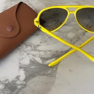 May include: Bright yellow sunglasses with dark lenses and a matching case. The sunglasses have a classic aviator shape with a double bridge and a curved brow bar. The case is brown and has a snap closure.