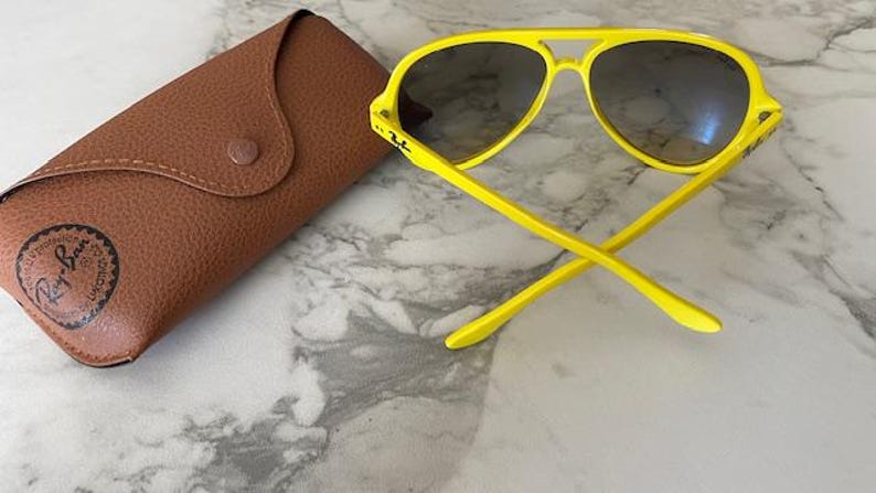 May include: Bright yellow sunglasses with dark lenses and a matching case. The sunglasses have a classic aviator shape with a double bridge and a curved brow bar. The case is brown and has a snap closure.
