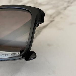 May include: Black sunglasses with dark tinted lenses. The sunglasses have a thick, square frame and the word "PRIZM" is visible on the lens. The sunglasses are resting on a light-colored surface.