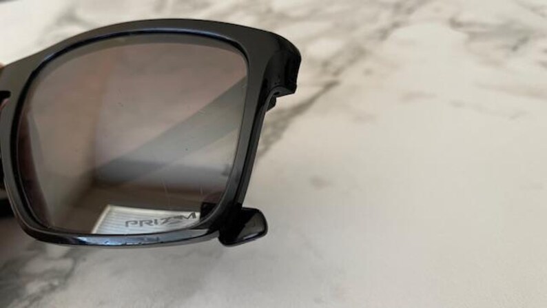 May include: Black sunglasses with dark tinted lenses. The sunglasses have a thick, square frame and the word "PRIZM" is visible on the lens. The sunglasses are resting on a light-colored surface.