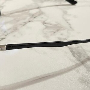 May include: Black sunglasses with dark lenses. The sunglasses have a sleek, modern design with a black frame and arms. The Ray-Ban logo is visible on the arm. The sunglasses are resting on a white and gray marble surface.