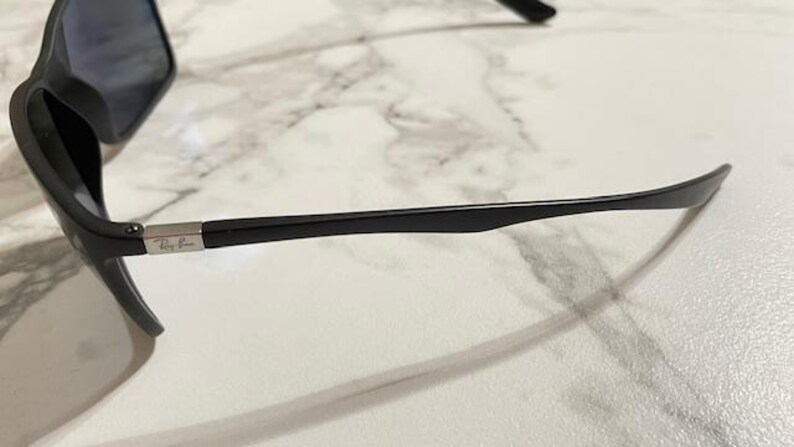 May include: Black sunglasses with dark lenses. The sunglasses have a sleek, modern design with a black frame and arms. The Ray-Ban logo is visible on the arm. The sunglasses are resting on a white and gray marble surface.