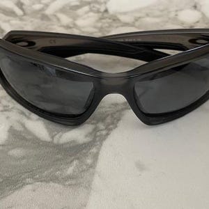 May include: Dark gray sunglasses with a wraparound design. The sunglasses have a thick frame and dark lenses. The glasses are resting on a light-colored, marble-like surface.