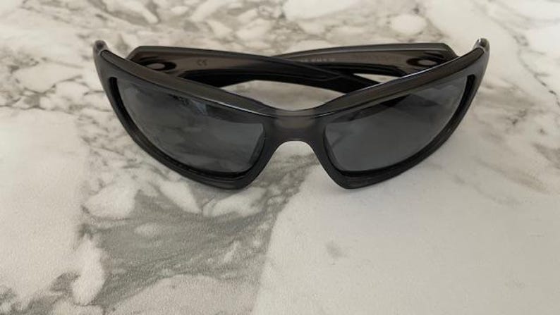 May include: Dark gray sunglasses with a wraparound design. The sunglasses have a thick frame and dark lenses. The glasses are resting on a light-colored, marble-like surface.