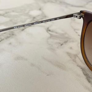 May include: Close-up of sunglasses with brown frames and gradient lenses. The arm of the glasses is silver with the text "RB4171 ERIKA 54&13 145 3N". The sunglasses are set against a marble background.