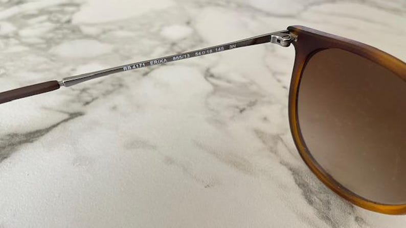 May include: Close-up of sunglasses with brown frames and gradient lenses. The arm of the glasses is silver with the text "RB4171 ERIKA 54&13 145 3N". The sunglasses are set against a marble background.