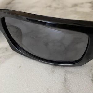 K&ouml;nnte beinhalten: Nahaufnahme einer schwarzen Sonnenbrille mit dunkelgrauen Gl&auml;sern. Die Sonnenbrille hat ein schlankes, modernes Design mit einem dicken Rahmen und gebogenen B&uuml;geln. Die Gl&auml;ser sind leicht reflektierend.
