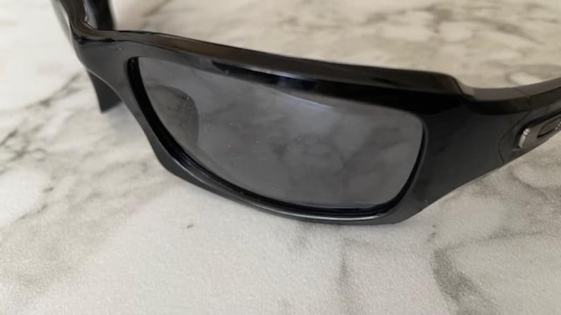 K&ouml;nnte beinhalten: Nahaufnahme einer schwarzen Sonnenbrille mit dunkelgrauen Gl&auml;sern. Die Sonnenbrille hat ein schlankes, modernes Design mit einem dicken Rahmen und gebogenen B&uuml;geln. Die Gl&auml;ser sind leicht reflektierend.