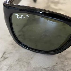 Puede incluir: Primer plano de gafas de sol negras con lentes verdes. El logotipo de Ray-Ban es visible en la parte superior izquierda de la lente. Las gafas de sol tienen un dise&ntilde;o elegante y curvado.
