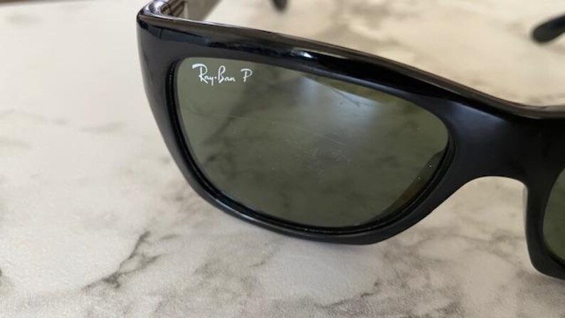 Puede incluir: Primer plano de gafas de sol negras con lentes verdes. El logotipo de Ray-Ban es visible en la parte superior izquierda de la lente. Las gafas de sol tienen un dise&ntilde;o elegante y curvado.