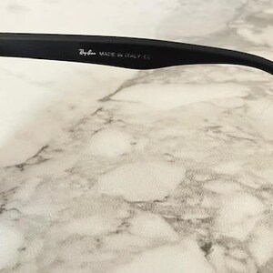 Pu&ograve; includere: Primo piano di occhiali da sole neri con lenti scure. Il logo Ray-Ban e "MADE IN ITALY CE" sono stampati sull'asta. Gli occhiali da sole sono appoggiati su una superficie simile al marmo.