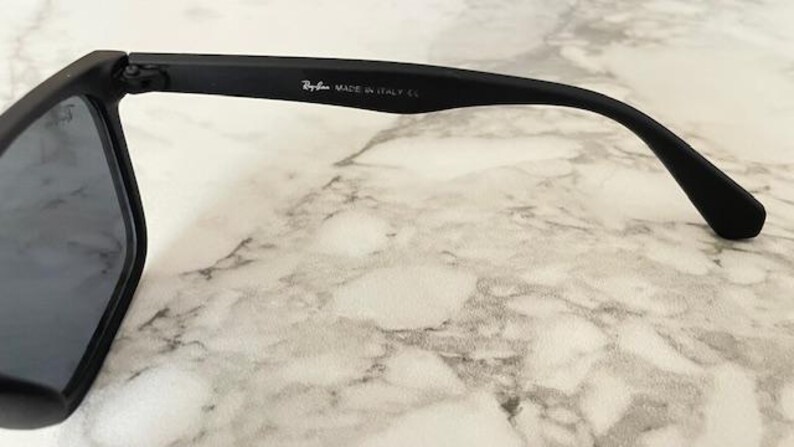 Pu&ograve; includere: Primo piano di occhiali da sole neri con lenti scure. Il logo Ray-Ban e "MADE IN ITALY CE" sono stampati sull'asta. Gli occhiali da sole sono appoggiati su una superficie simile al marmo.