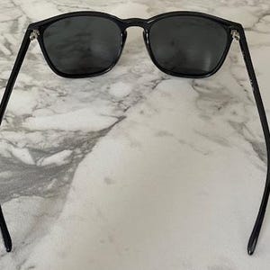 Puede incluir: Gafas de sol negras con lentes oscuras y montura redondeada. Las gafas tienen patillas finas y negras que se curvan hacia afuera. Las gafas est&aacute;n sobre un fondo de m&aacute;rmol.