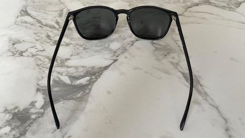 Puede incluir: Gafas de sol negras con lentes oscuras y montura redondeada. Las gafas tienen patillas finas y negras que se curvan hacia afuera. Las gafas est&aacute;n sobre un fondo de m&aacute;rmol.
