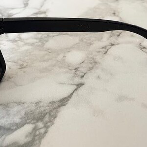May include: Black sunglasses with gray lenses. The frame is thick and angular. The temple arm has a red and white "USA" logo. The sunglasses are set against a white and gray marble background.