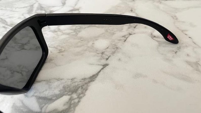 May include: Black sunglasses with gray lenses. The frame is thick and angular. The temple arm has a red and white "USA" logo. The sunglasses are set against a white and gray marble background.